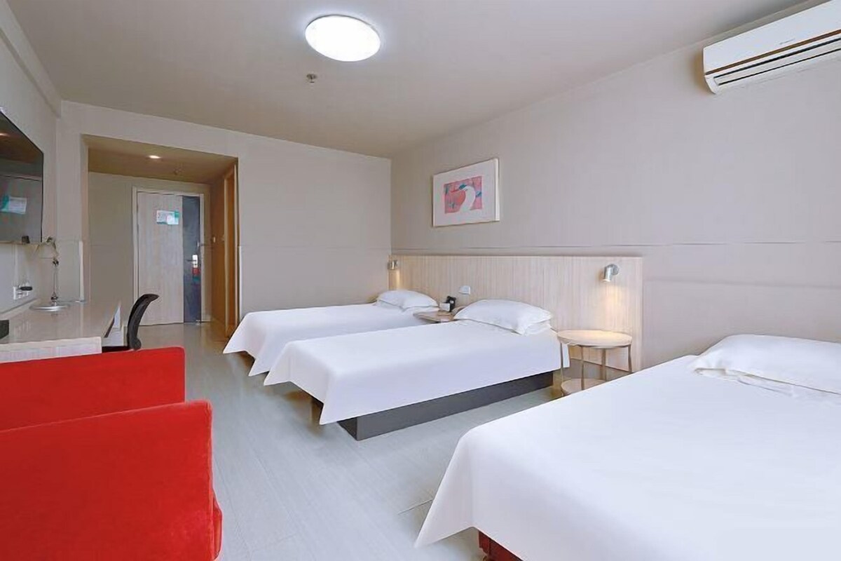 A hotel room with three beds, a red chair, a desk with a lamp, and an air conditioning unit.