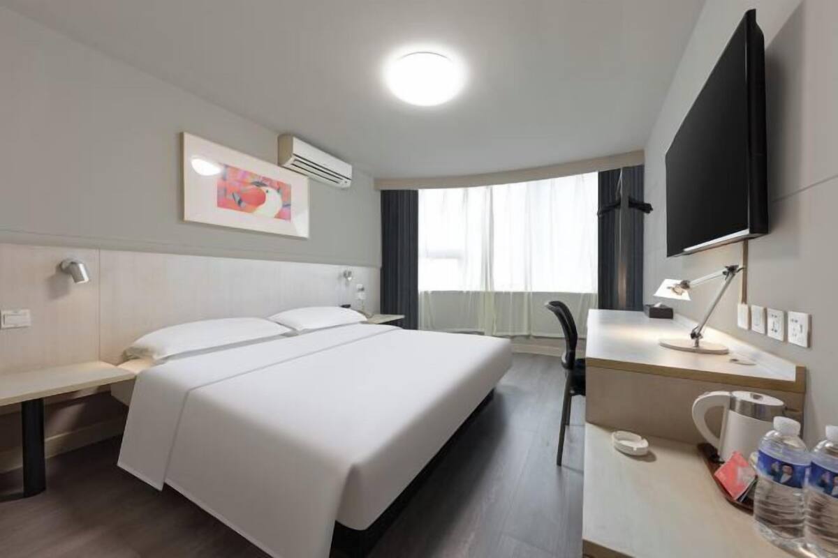 A hotel room with a bed, bedside table, desk, chair, flat-screen TV, air conditioning unit, and a window with curtains.