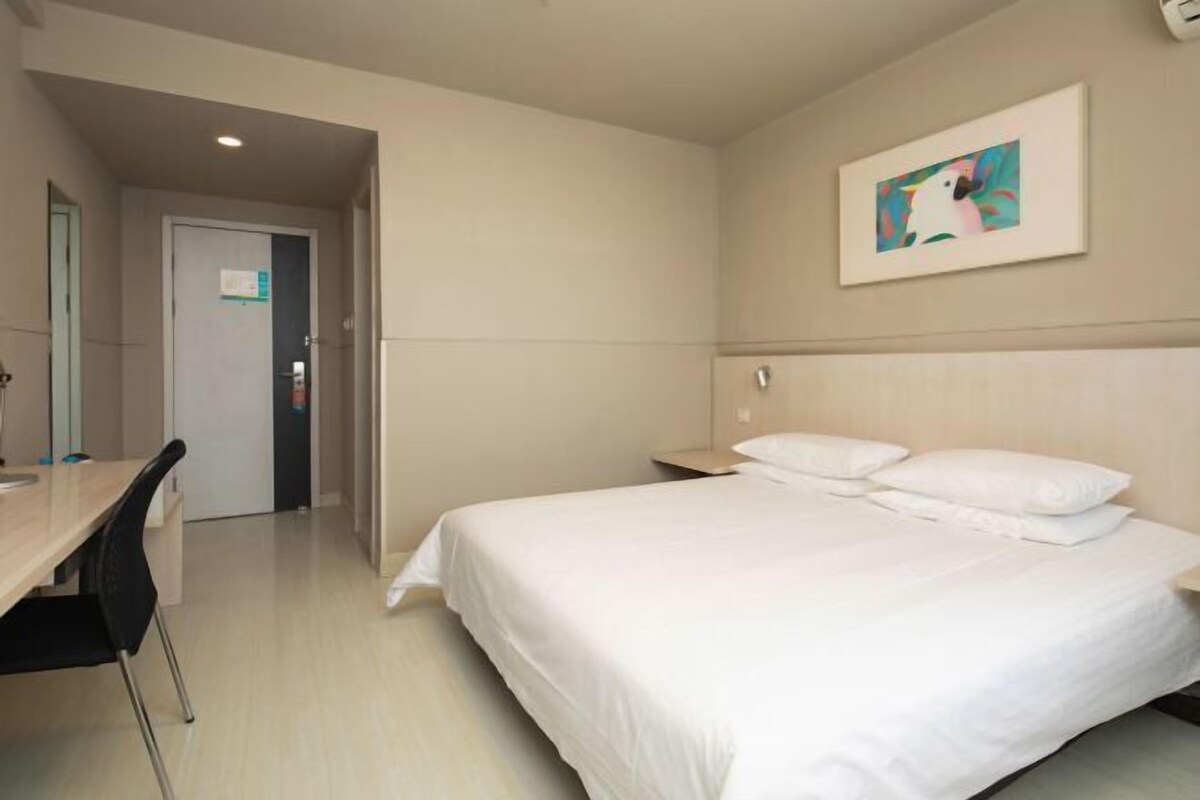 A hotel room with a bed, a desk, and a chair. There is a door leading to another room and a painting on the wall.