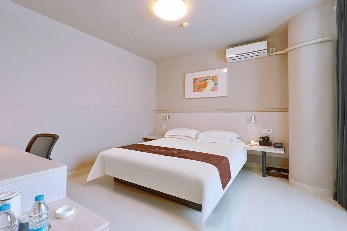 A hotel room with a bed, bedside table, desk, chair, and an air conditioning unit.