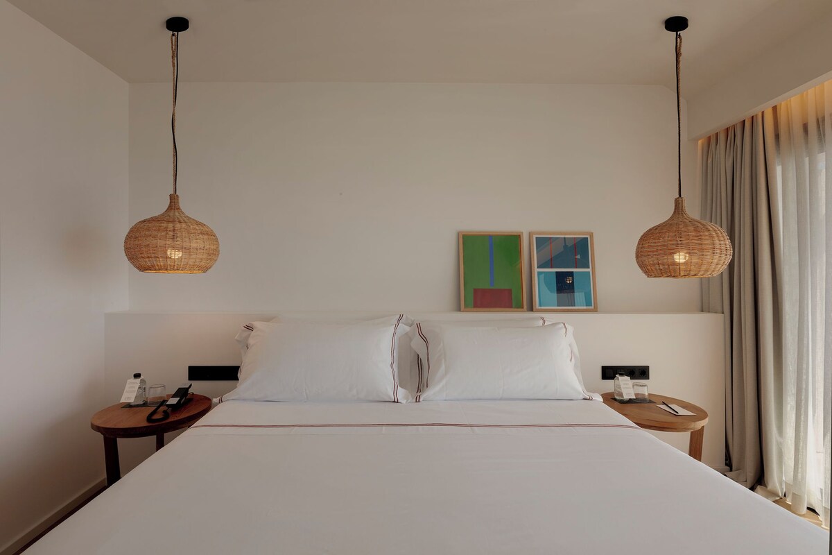 Habitación de hotel con una cama grande, dos mesitas de noche, una lámpara colgante y dos cuadros enmarcados en la pared.