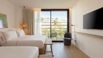 Habitación de hotel con dos camas, un ventanal amplio con vistas a edificios, un televisor de pantalla plana montado en la pared y una mesita con una silla.
