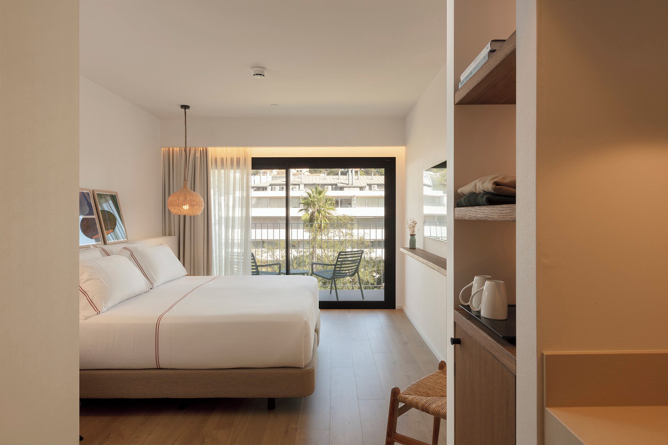 A bedroom with a bed, a chair, a shelf with cups, and a balcony with a view of buildings and a palm tree.