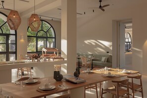 Une salle à manger spacieuse, avec une longue table en bois dressée pour le repas, entourée de chaises en osier. De grandes fenêtres laissent entrer la lumière naturelle et offrent une vue sur la verdure environnante.
