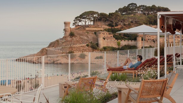 Una terraza frente al mar con sillas de mimbre y una mesa, con vistas a un fuerte histórico y al océano.
