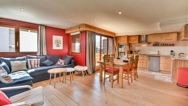 A modern living room with a sofa, dining table, and chairs. There is a kitchen area with wooden cabinets and appliances.