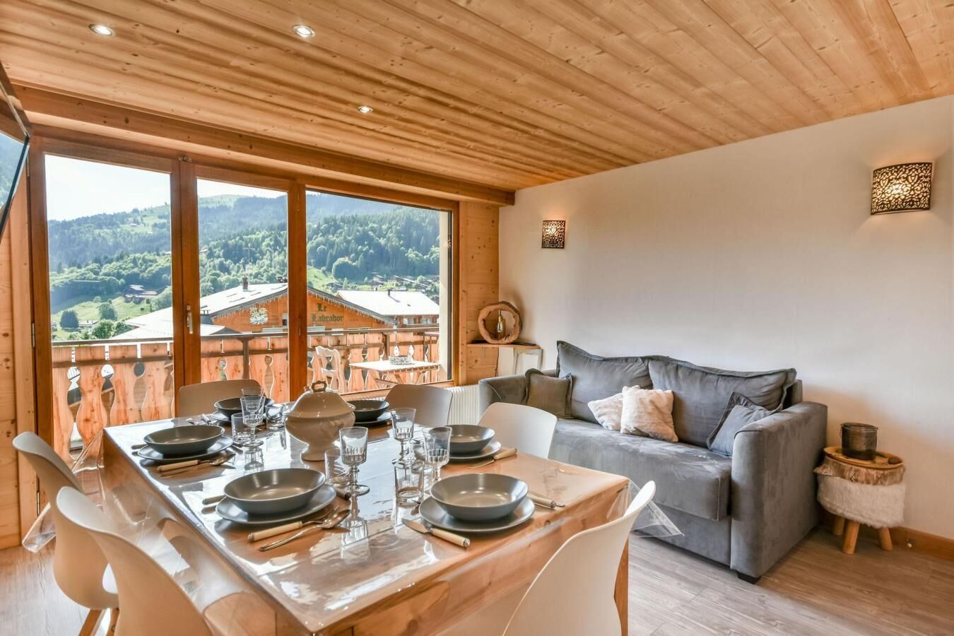 Un comedor con una mesa de madera dispuesta para cuatro personas, un sofá gris y vistas a un paisaje montañoso a través de ventanas grandes.