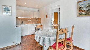 Een kleine keuken met witte kastjes, een koelkast en een houten tafel met een bloemrijk tafelkleed.