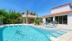A swimming pool with clear water, surrounded by a patio area with lounge chairs and a building with a tiled roof.