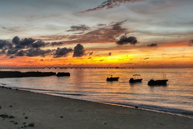 A sunset over a calm sea with boats and a pier in the distance.