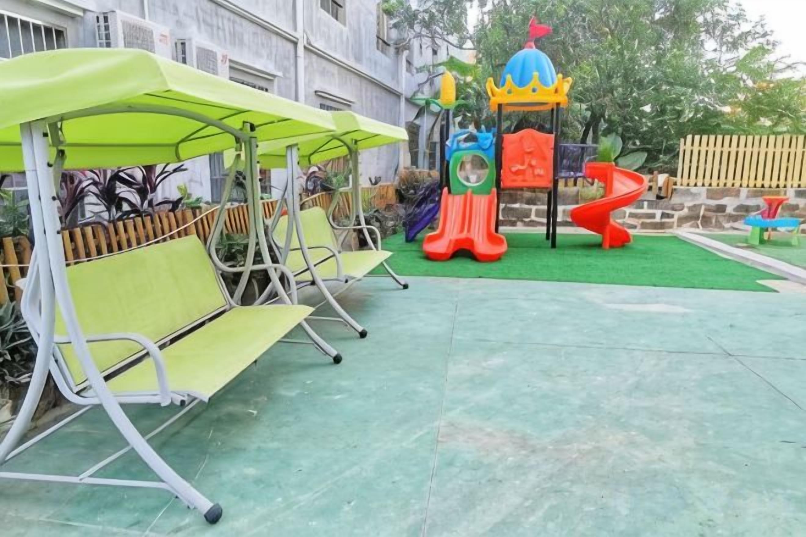 A playground with slides and a climbing structure, surrounded by green artificial grass and metal benches.