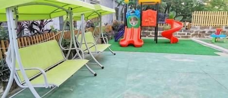 A playground with slides and a climbing structure, surrounded by green artificial grass and metal benches.