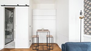 Huoneessa on kalanruotokuviollinen puulattia, sininen samettituoli, pieni pöytä ja kaksi jakkaraa sekä kuvioitu tapetti seinällä.
