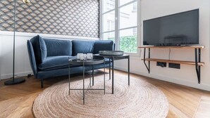 Moderni olohuone, jossa on sininen sohva, pyöreä, kudottu matto, litteänäyttöinen televisio puisen jalustan päällä ja kuvioitu seinä.