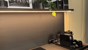 Un comptoir de cuisine avec une machine à café, une bouilloire et un distributeur de thé. Un tableau encadré est accroché au mur, au-dessus du comptoir.