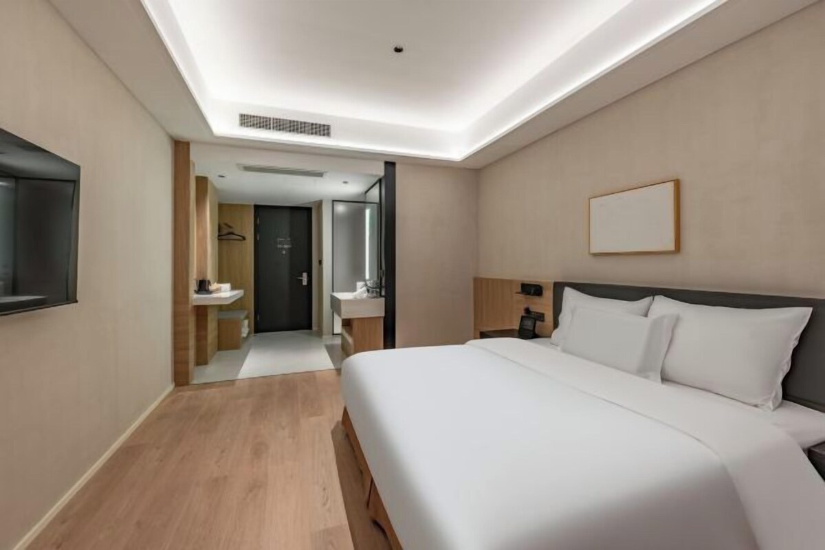 A hotel room with a large bed, a flat-screen TV, a bedside table, and a bathroom with a sink and mirror.