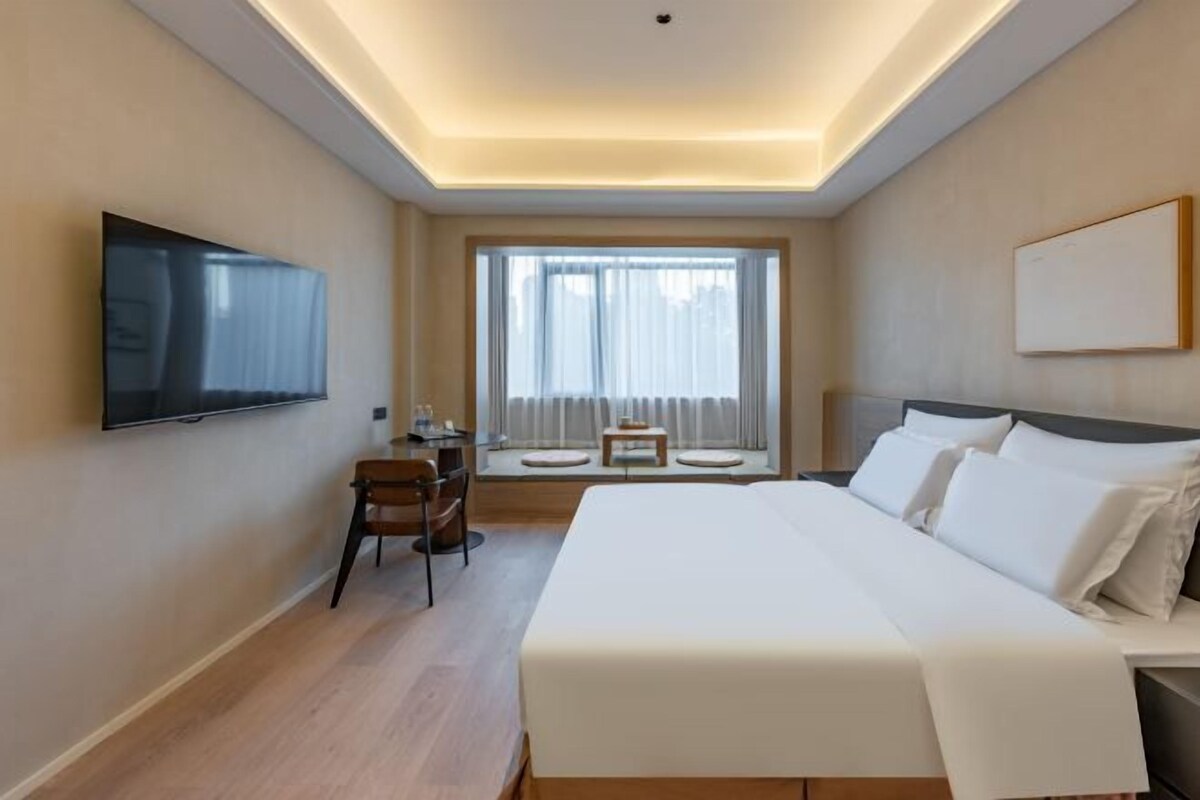 A hotel room with a large bed, a flat-screen TV mounted on the wall, a desk with a chair, and a window with curtains.