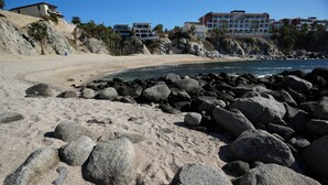 Ein Strand mit großen Felsen im Vordergrund, ein sandiger Strand und Gebäude im Hintergrund.