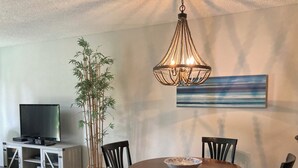 A dining area with a wooden table, chairs, a bowl, a television, a bamboo plant, and a chandelier. - STEPS TO THE BEACH and POOL Oceanfront QUIET complex Lola’sLanding!! CouplesOnly (St. Augustine)