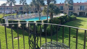 A pool area with a clear view of the pool, surrounded by a white fence, palm trees, and a building with balconies in the background. - STEPS TO THE BEACH and POOL Oceanfront QUIET complex Lola’sLanding!! CouplesOnly (St. Augustine)