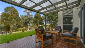 A covered outdoor deck with wooden furniture and a glass ceiling, surrounded by a well-maintained garden and trees.