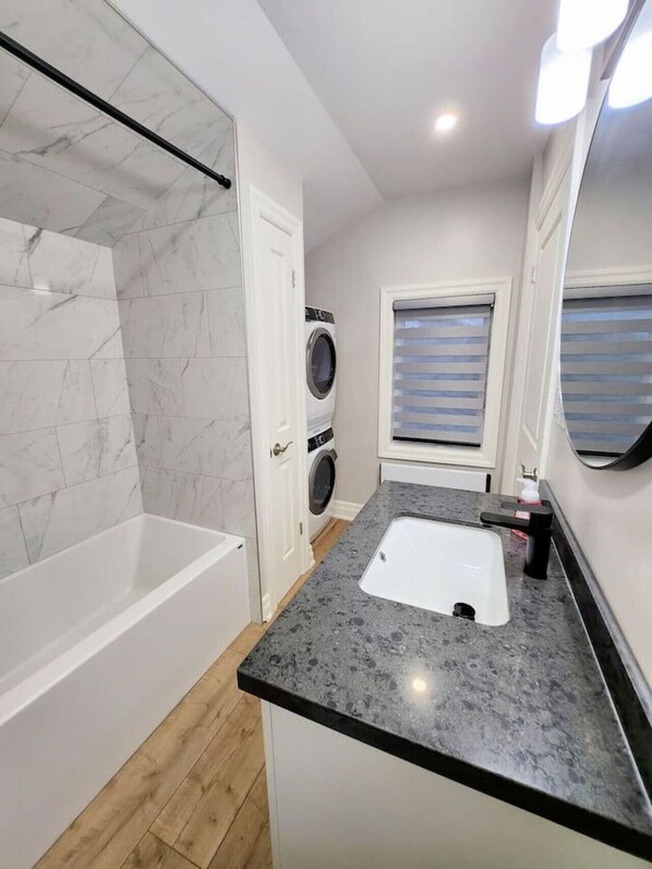 A modern bathroom with a marble shower, a built-in washing machine, a sink with a countertop, and a window with blinds.