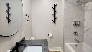 A modern bathroom with a marble shower, a sink with a black faucet, and a mirror with a black leaf-shaped decoration. - The Cozy Cove Modern King-Bedroom Full Apartment (St. Catharines)
