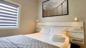 A hotel room with a bed, a window with blinds, a framed picture on the wall, and a nightstand with a lamp. - The Zen Escape Modern Queen-Bed Full Apartment (St. Catharines)