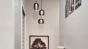 A hallway with wooden flooring, a framed tree artwork, pendant lighting, and a white pedestal. - The Zen Escape Modern Queen-Bed Full Apartment (St. Catharines)