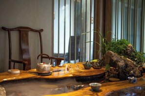 A wooden table with a teapot, cups, and a small bowl, set against a backdrop of a wooden chair and a window with vertical blinds. - Lijiang Shechen Inn (Lijiang)