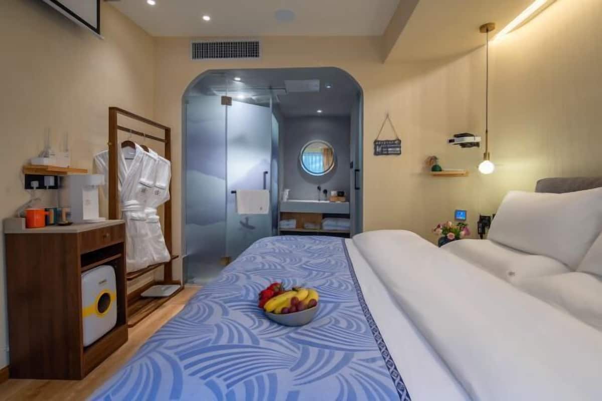 A hotel room with a bed, a bedside table with a bowl of fruit, a robe rack, and a small desk with a lamp.