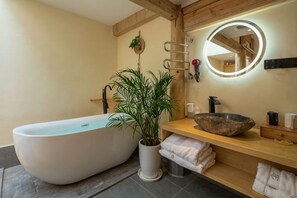 Ein modernes Badezimmer mit freistehender Badewanne, einem Topf mit Pflanze, einem hölzernen Waschtisch mit rundem Spiegel und einem Handtuchhalter.