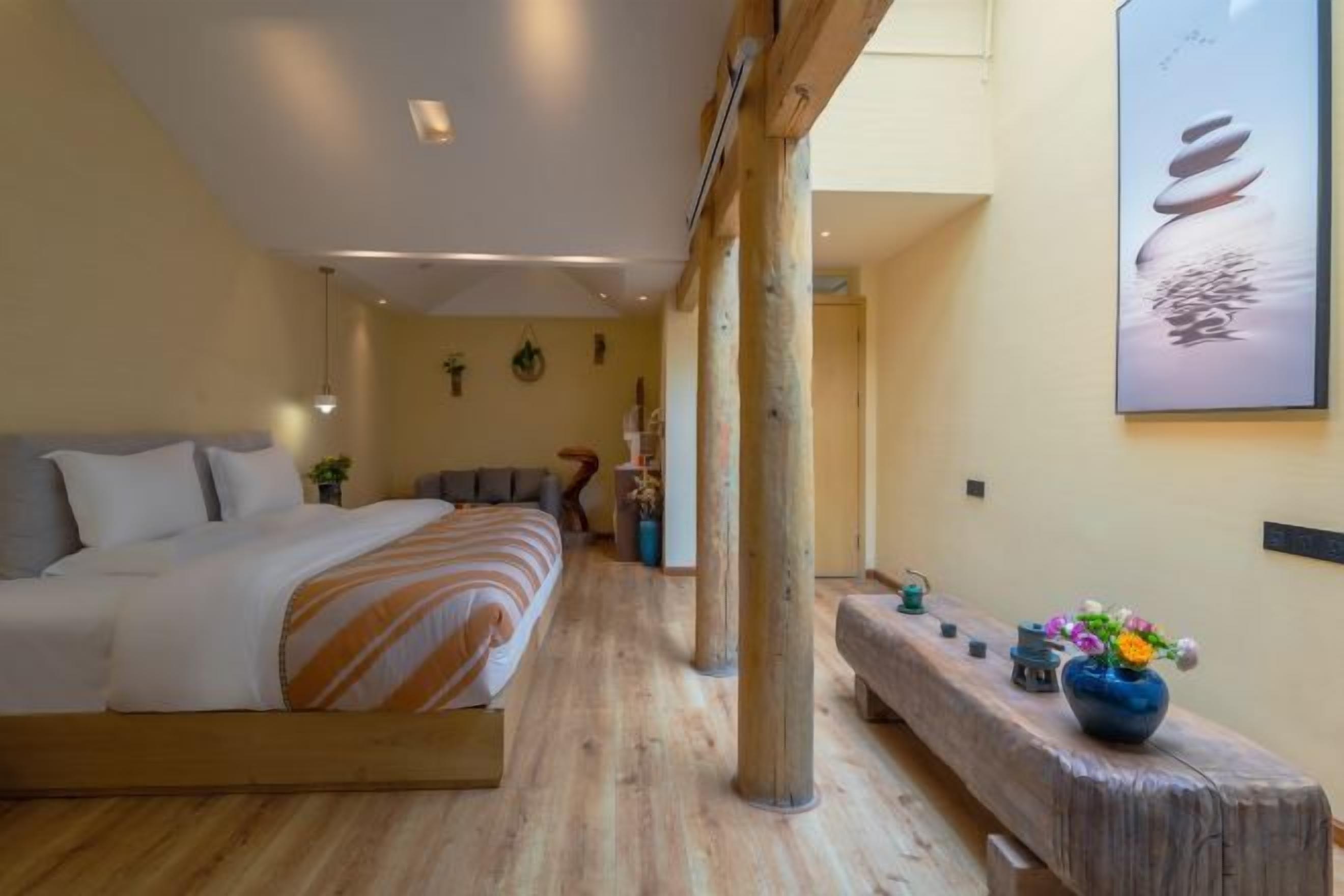 A bedroom with a bed, wooden floor, a bench, a vase with flowers, and a wall-mounted artwork.