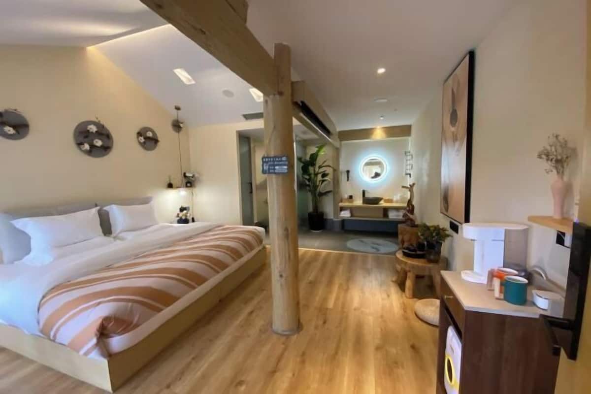 A hotel room with a bed, wooden floor, a small desk with a chair, a mirror, and a plant.