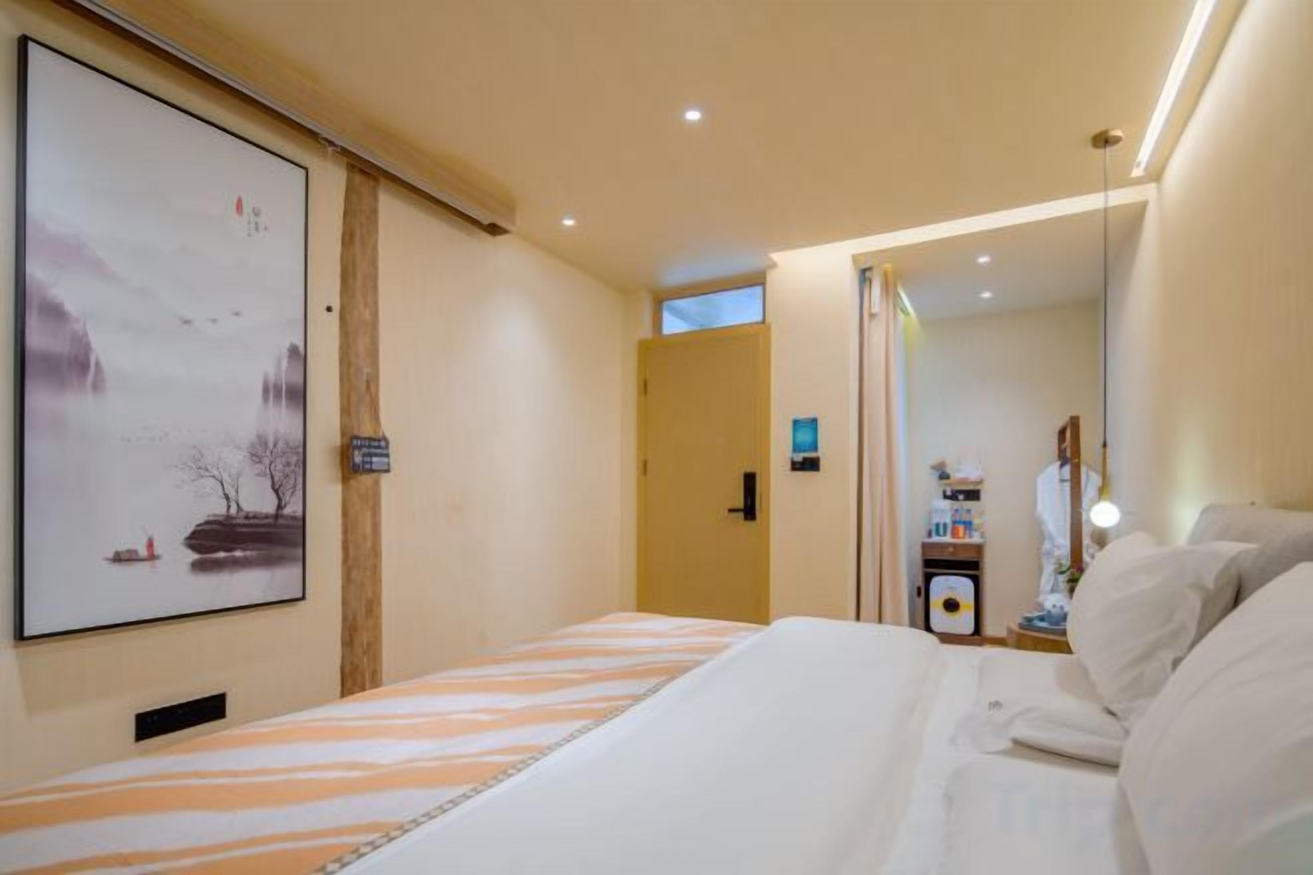 A hotel room with a bed, a painting, a door, and a small table with a lamp.