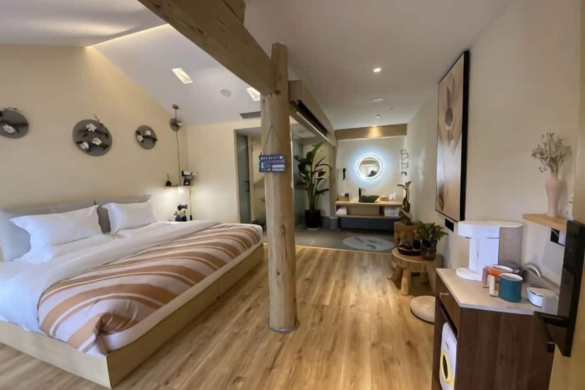 A modern hotel room with a large bed, wooden flooring, a desk with a chair, and a bathroom visible in the background.