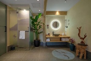 Ein modernes Badezimmer mit Duschkabine aus Glas, einem runden Spiegel, einem Waschbecken mit Schale, einer Topfpflanze und einer Holzskulptur.