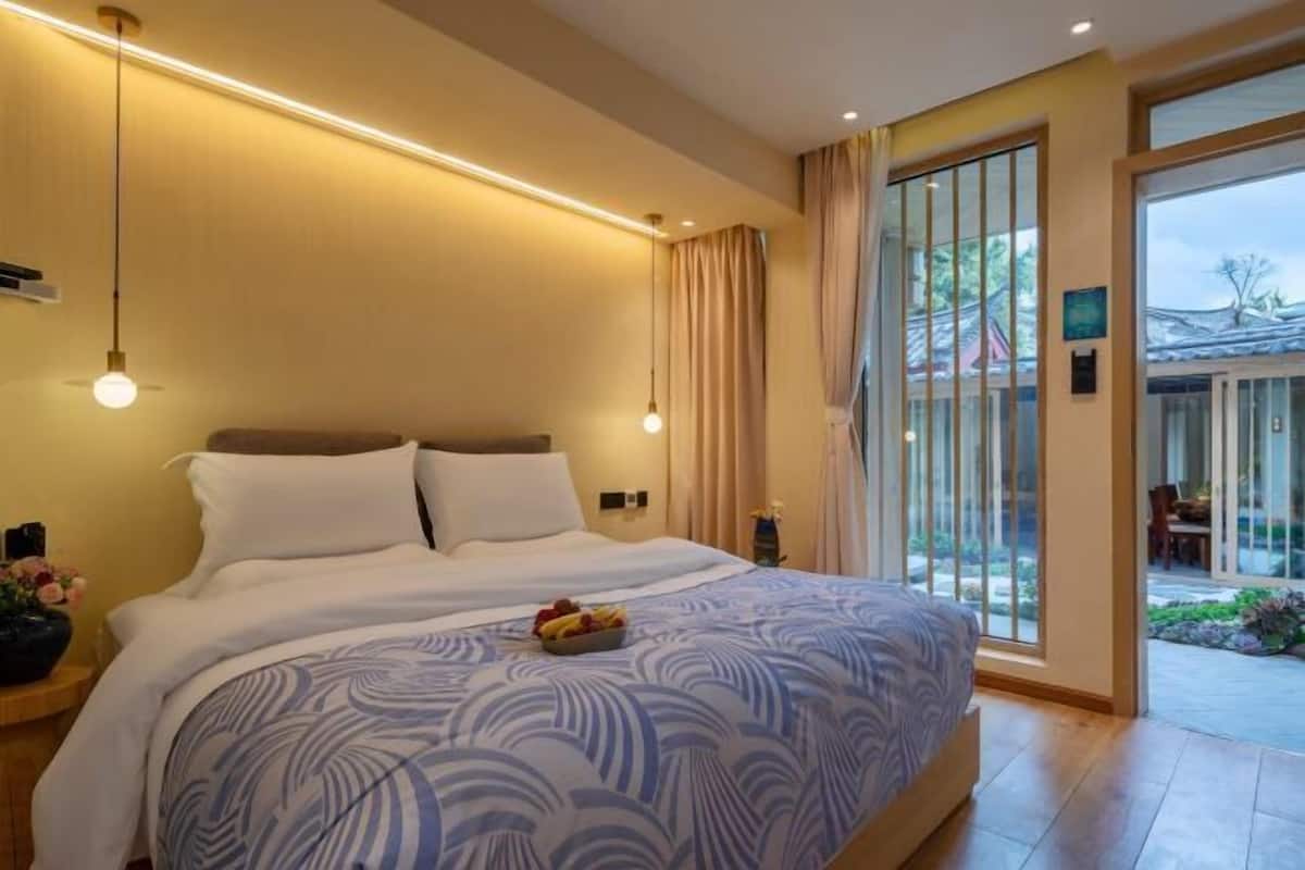 A modern hotel room with a large bed, a bedside table with a lamp, and a view of the outdoor garden through a sliding glass door.