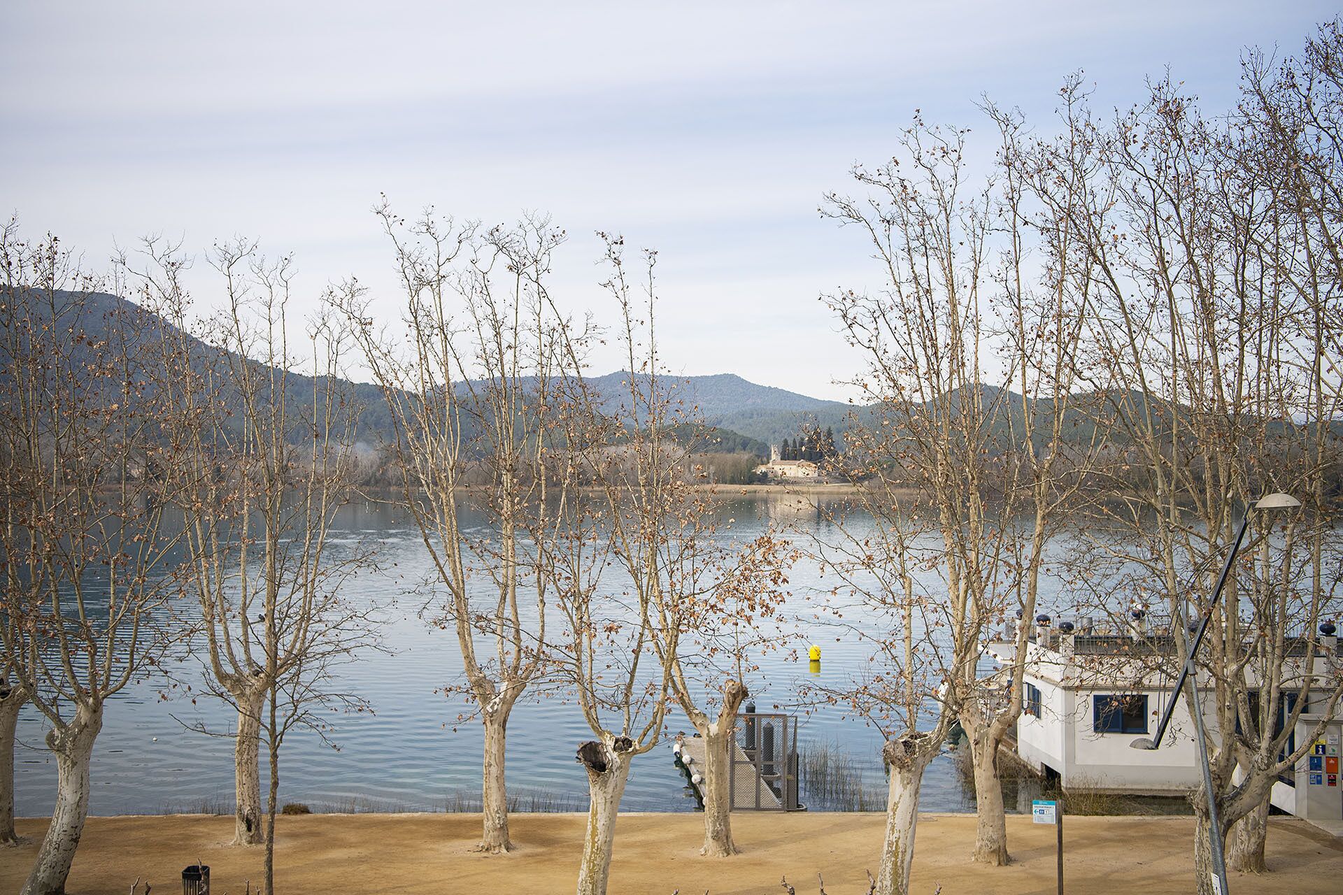 Vista de un lago con árboles desprovistos de hojas, un edificio lejano y un muelle.