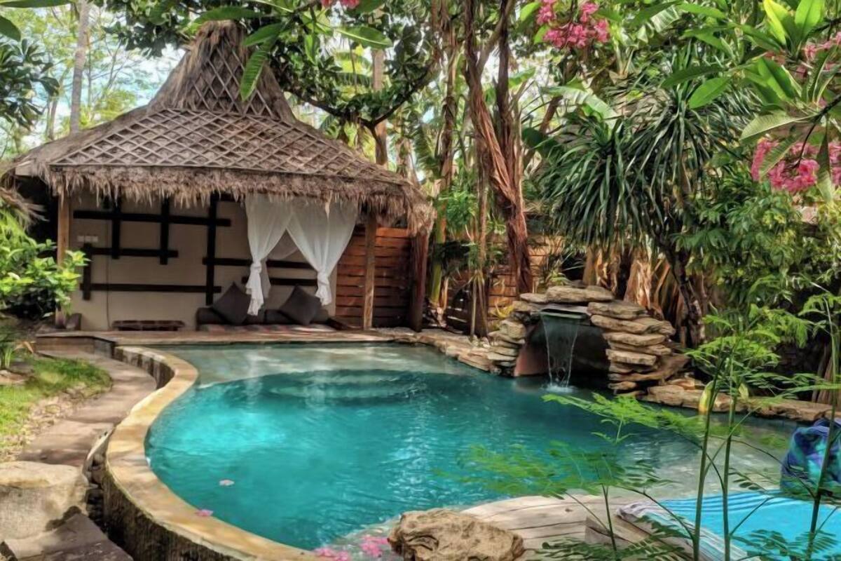 Un espace piscine agrémenté d’un petit pavillon à toit de chaume, entouré d’une végétation luxuriante et d’une petite cascade.