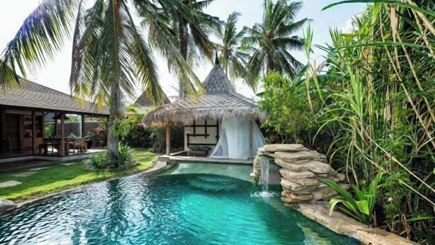 A pool with a waterfall, surrounded by lush greenery and a thatched-roof structure. - Villa Bhuvana with Private Swimming Pool (Gili Trawangan)