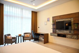 Une chambre d’hôtel comprenant un lit, un bureau, deux chaises, un téléviseur fixé à un mur en marbre et une grande fenêtre avec des rideaux.