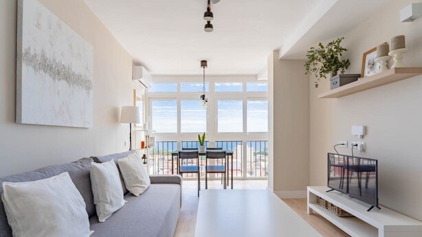Una sala moderna con un sofá gris, cojines blancos, una mesa de centro blanca y un televisor sobre un mueble blanco. Hay un balcón con una mesa y sillas, y un amplio ventanal que ofrece vistas de la ciudad.