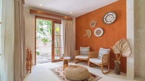 A room with a large window, two wicker chairs, a round ottoman, and decorative wall art. - White Goose Boutique Hotel (Bali)
