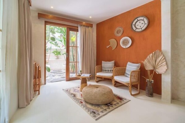 A room with a large window, two wicker chairs, a round ottoman, and decorative wall art. - White Goose Boutique Hotel (Bali)