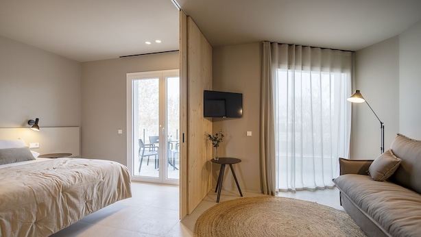 Quarto de hotel com cama, sofá, televisão, abajur e vista através de uma porta de vidro.
