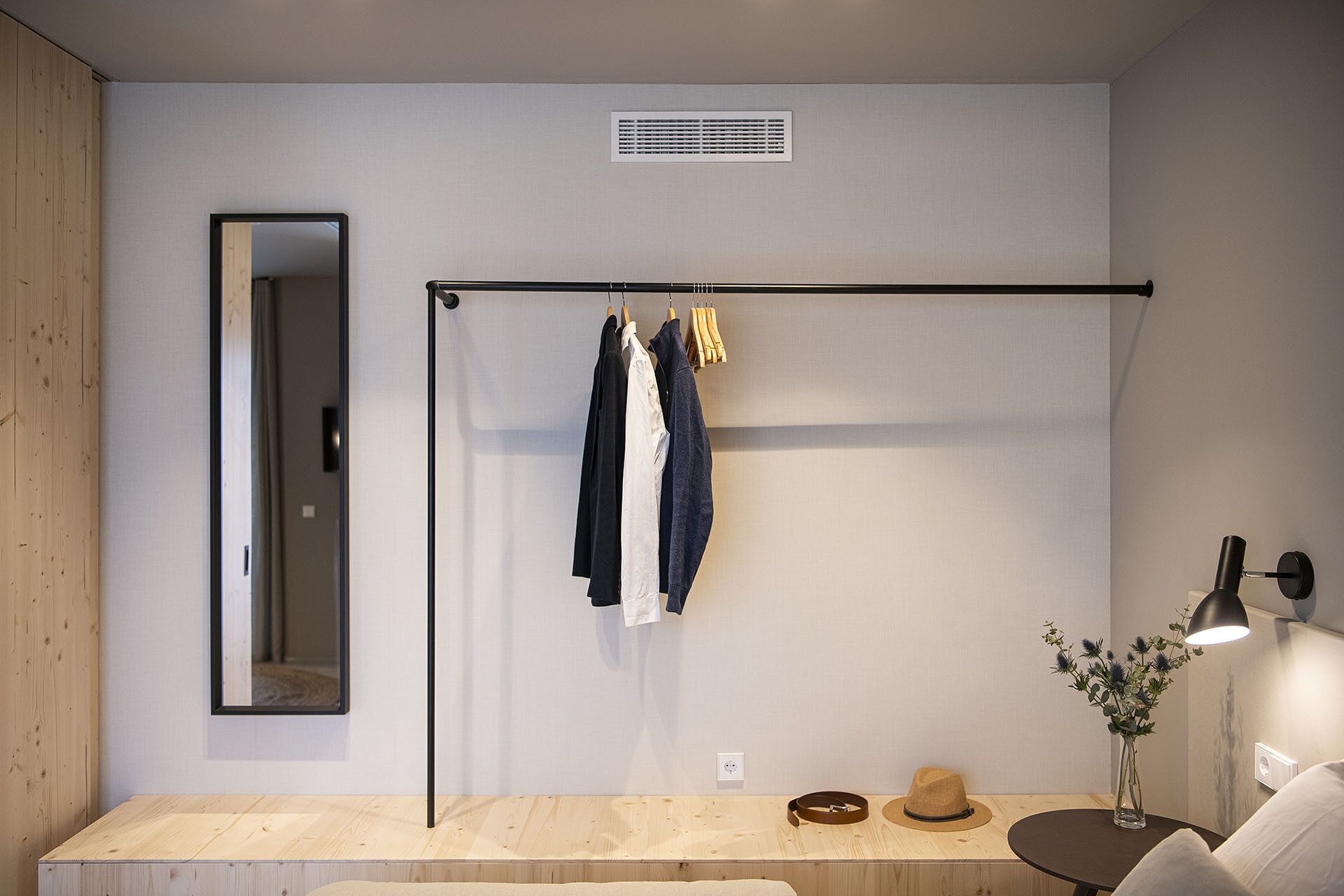 Quarto moderno com piso de madeira, uma estante de metal preta com roupas, um espelho, uma pequena mesa redonda com abajur e um vaso com flores.