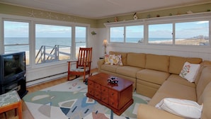 Una sala de estar con sofá, sillón, mesa de centro y televisión. Tiene ventanas amplias con vista a la playa y al océano.