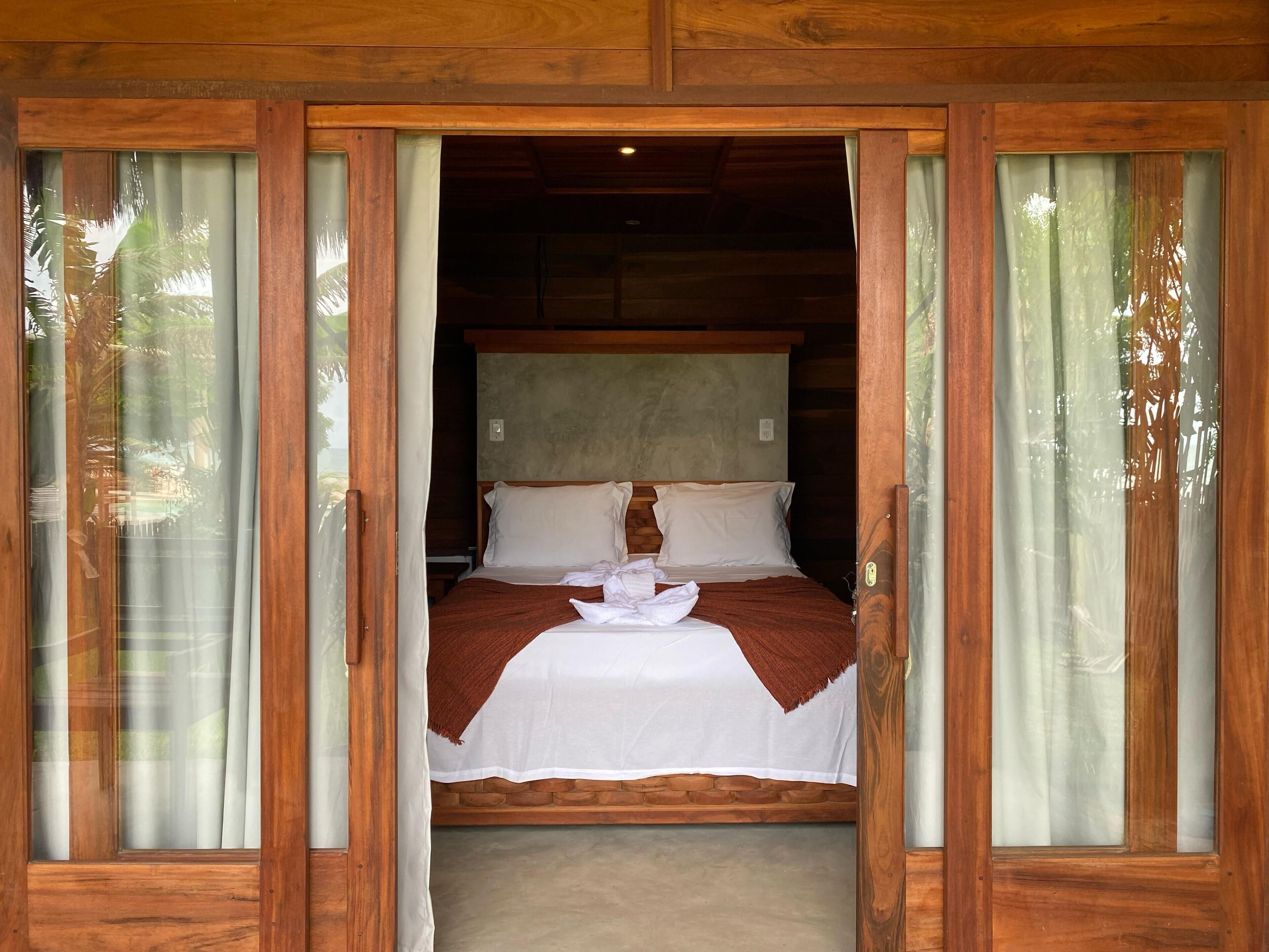 A wooden-framed room with a bed, curtains, and a view of greenery outside.