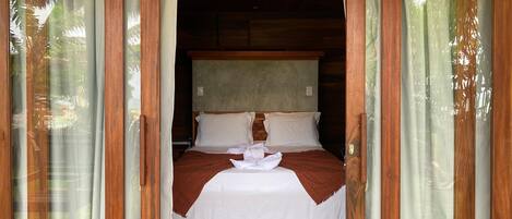 A wooden-framed room with a bed, curtains, and a view of greenery outside.